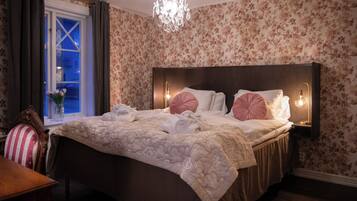 A hotel room with a large bed, floral wallpaper, a chandelier, and a window with curtains.