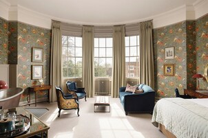A room with a large window, a bed, a blue armchair, a wooden side table, and a mirror.