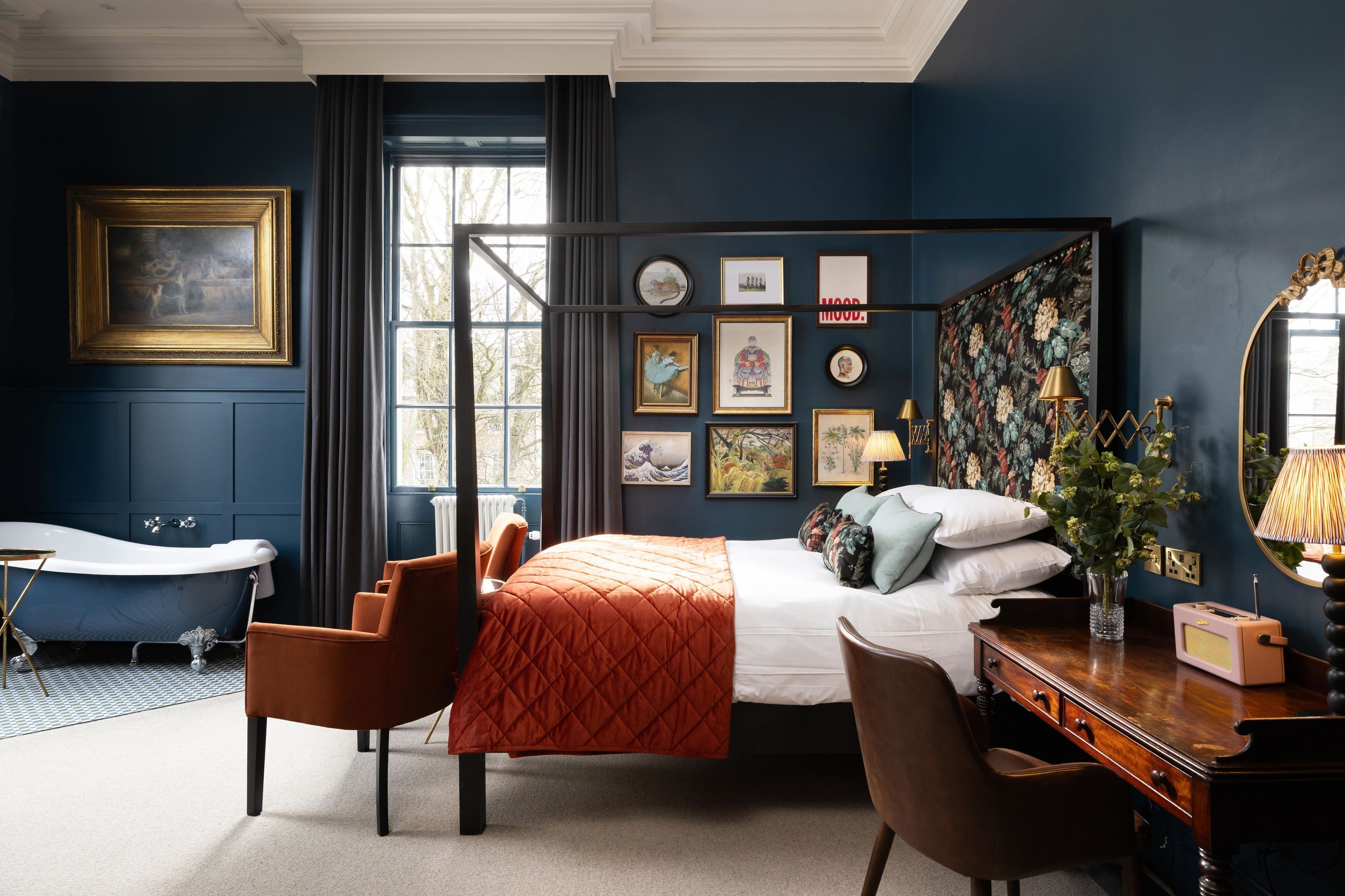 A bedroom with a large bed, a classic bathtub, a desk with a radio, and a mirror.
