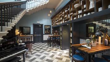 A grand piano in a spacious room with a checkered floor, a staircase, and a bar area with stools and a wooden table.