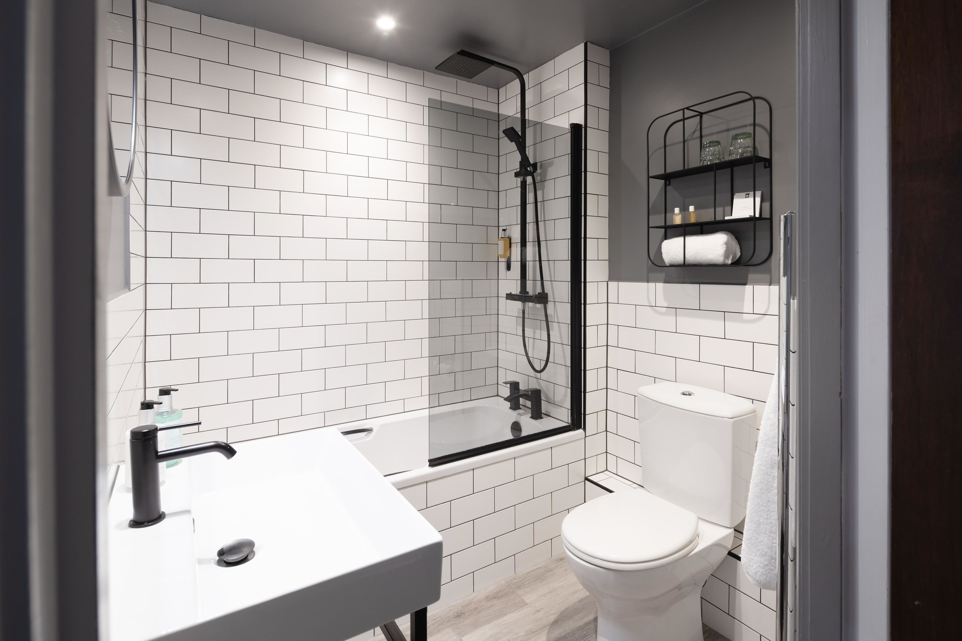Un baño moderno con paredes de azulejos blancos, cabina de ducha con mampara de vidrio, lavamanos blanco y un inodoro.