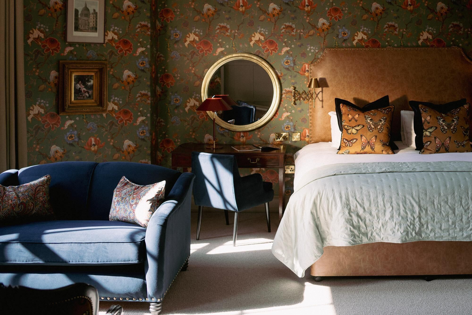 A bedroom with a floral wallpaper, a round mirror, a blue armchair, a wooden nightstand, and a bed with patterned pillows.