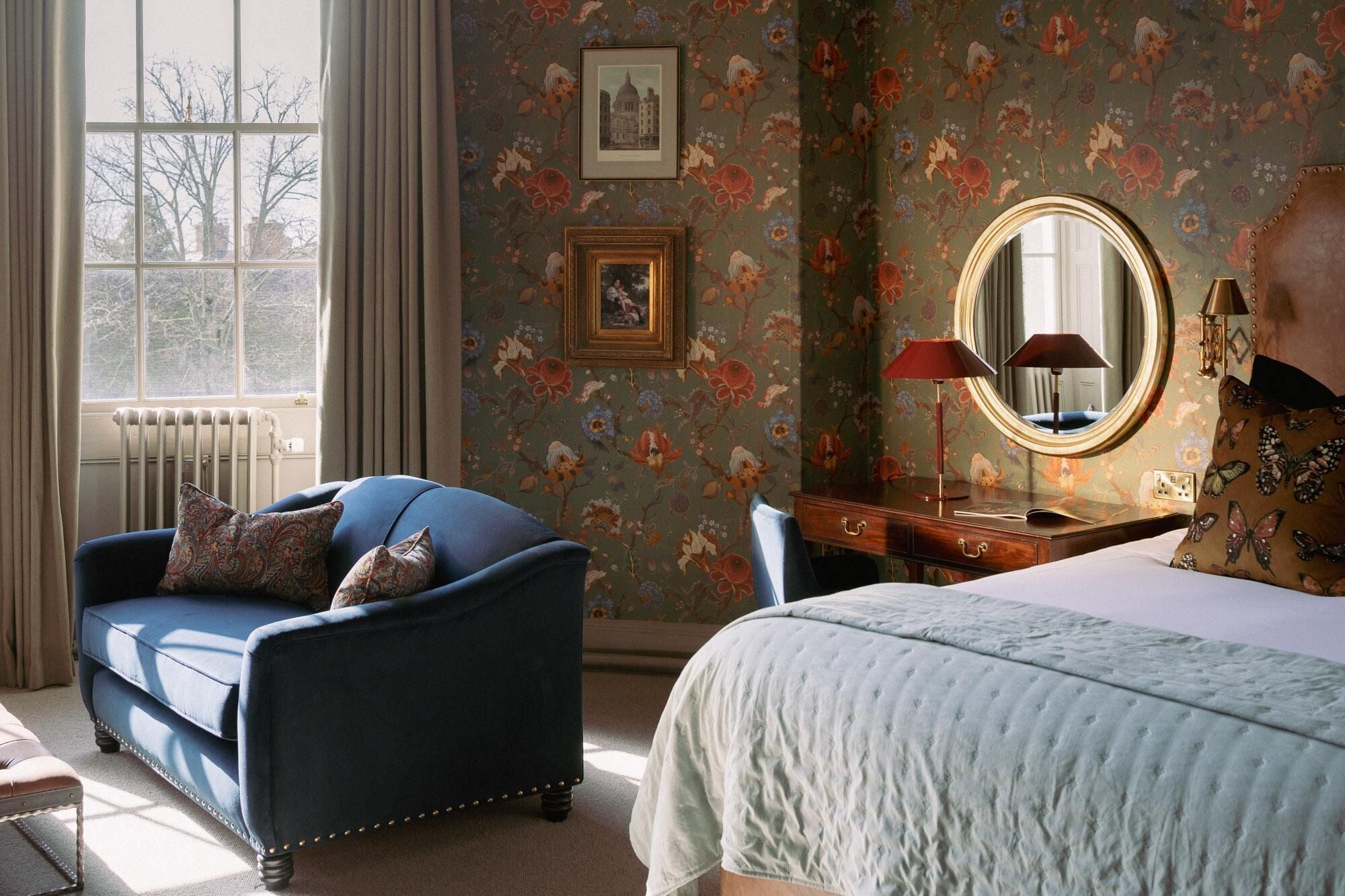 A room with a blue armchair, a bed with a white quilt, a wooden dresser, a round mirror, and floral wallpaper.