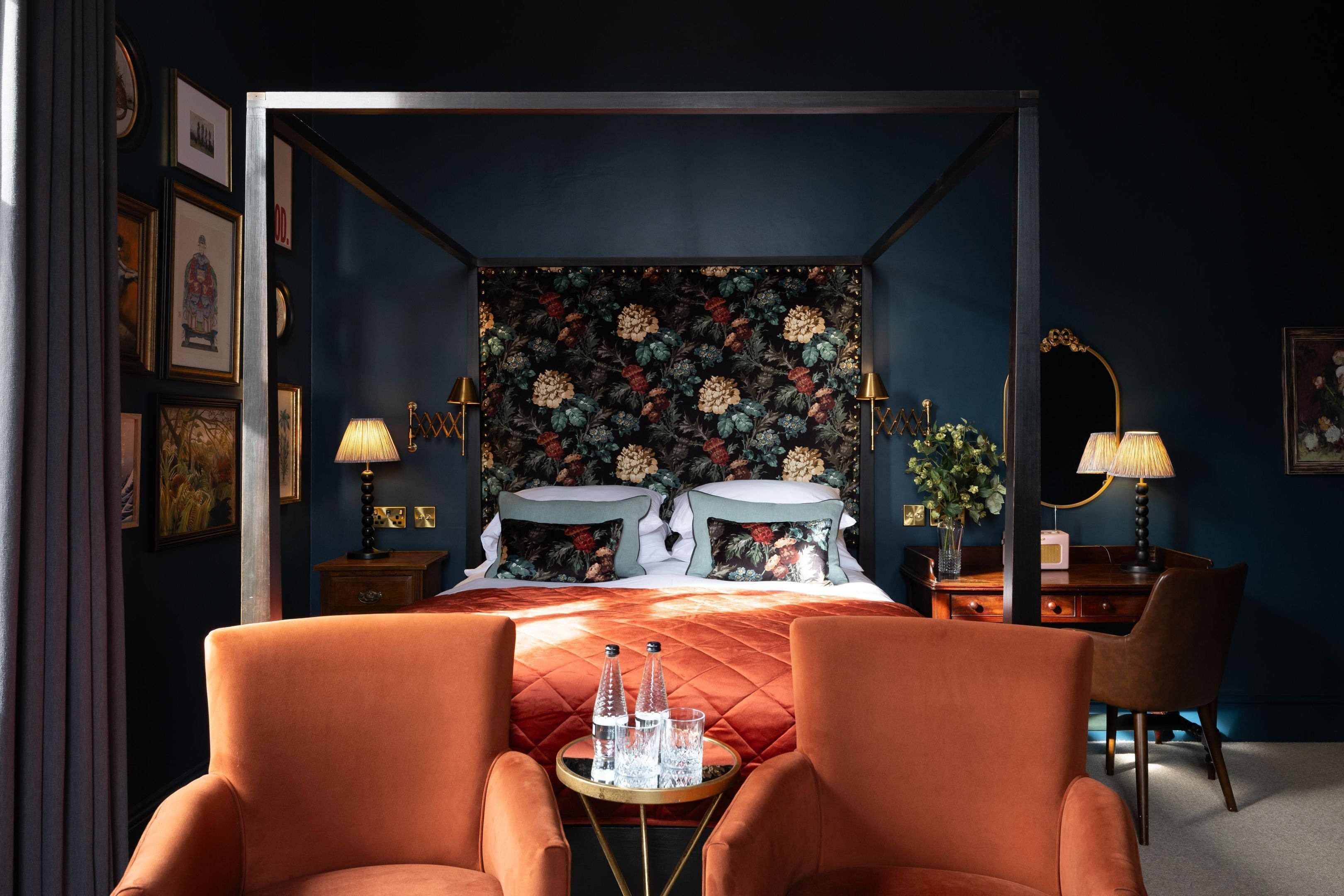 A four-poster bed with a floral headboard, two bedside tables with lamps, a desk with a mirror, and two armchairs.
