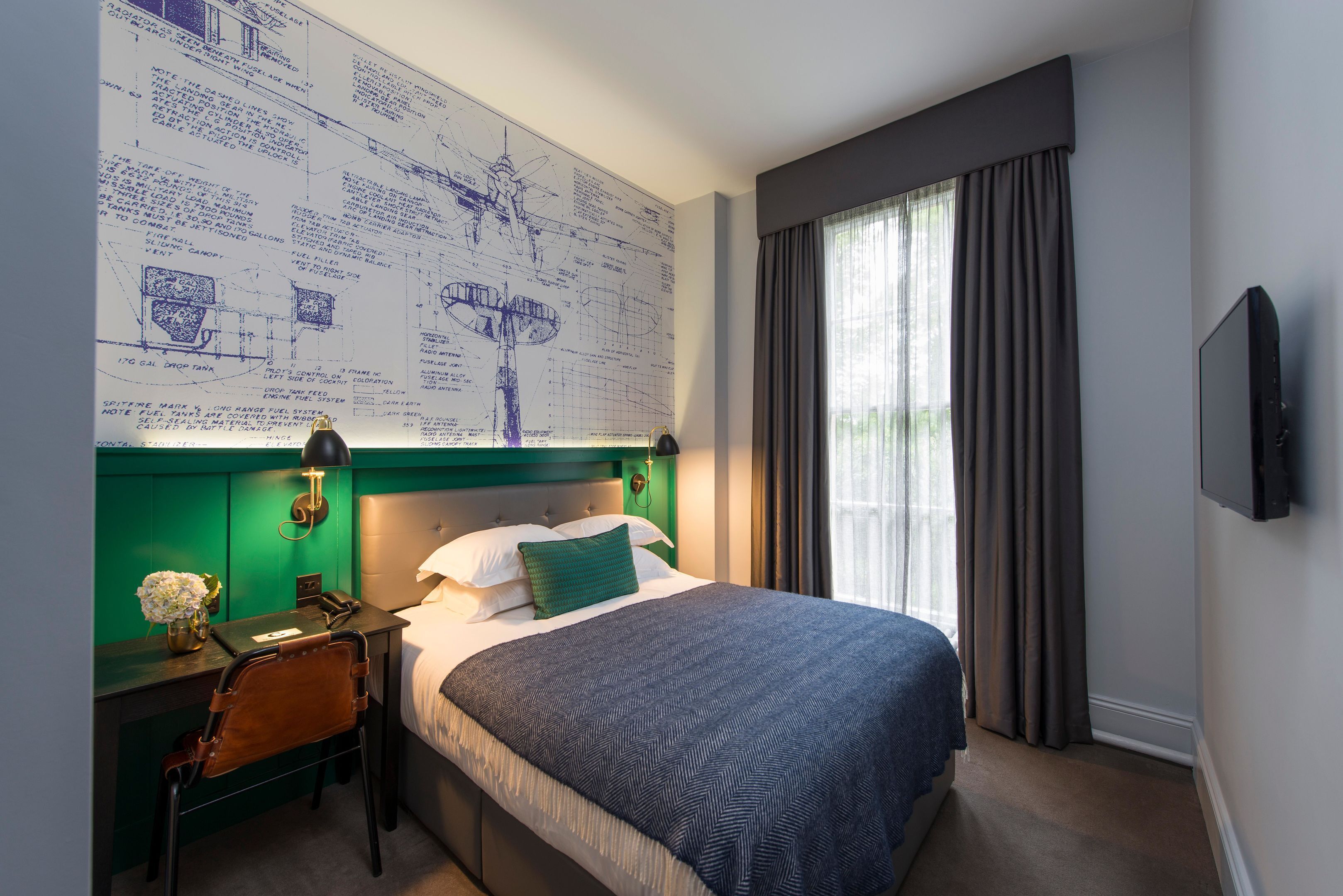 Ein Hotelzimmer mit einem großen Bett, einem Schreibtisch mit Stuhl, einem Nachttisch, einem an der Wand montierten Fernseher und einem Fenster mit Vorhängen.
