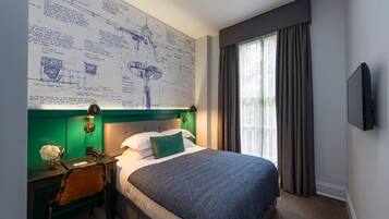 Ein Hotelzimmer mit einem großen Bett, einem Schreibtisch mit Stuhl, einem Nachttisch, einem an der Wand montierten Fernseher und einem Fenster mit Vorhängen.