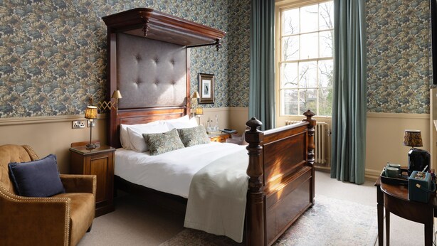 Un dormitorio con una cama grande, un cabecero de madera, una mesita de noche, una silla y una ventana con cortinas.