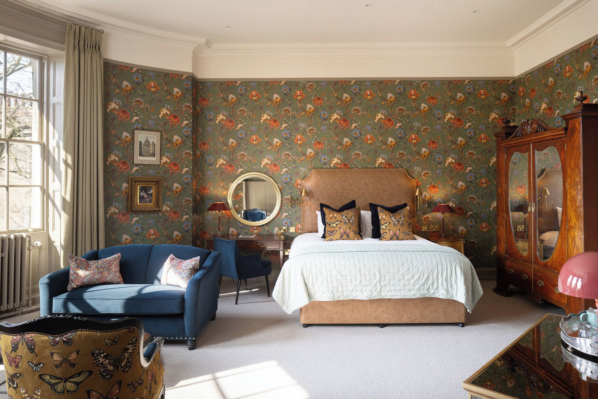 A bedroom with a floral wallpaper, a bed with patterned pillows, a blue armchair, and a wooden cabinet.