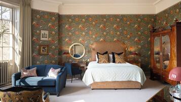 A bedroom with a floral wallpaper, a bed with patterned pillows, a blue armchair, and a wooden cabinet.