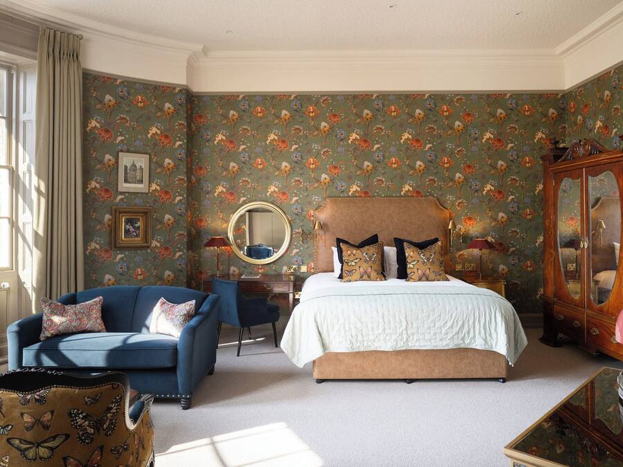 A bedroom with a floral wallpaper, a bed with patterned pillows, a blue armchair, and a wooden cabinet.