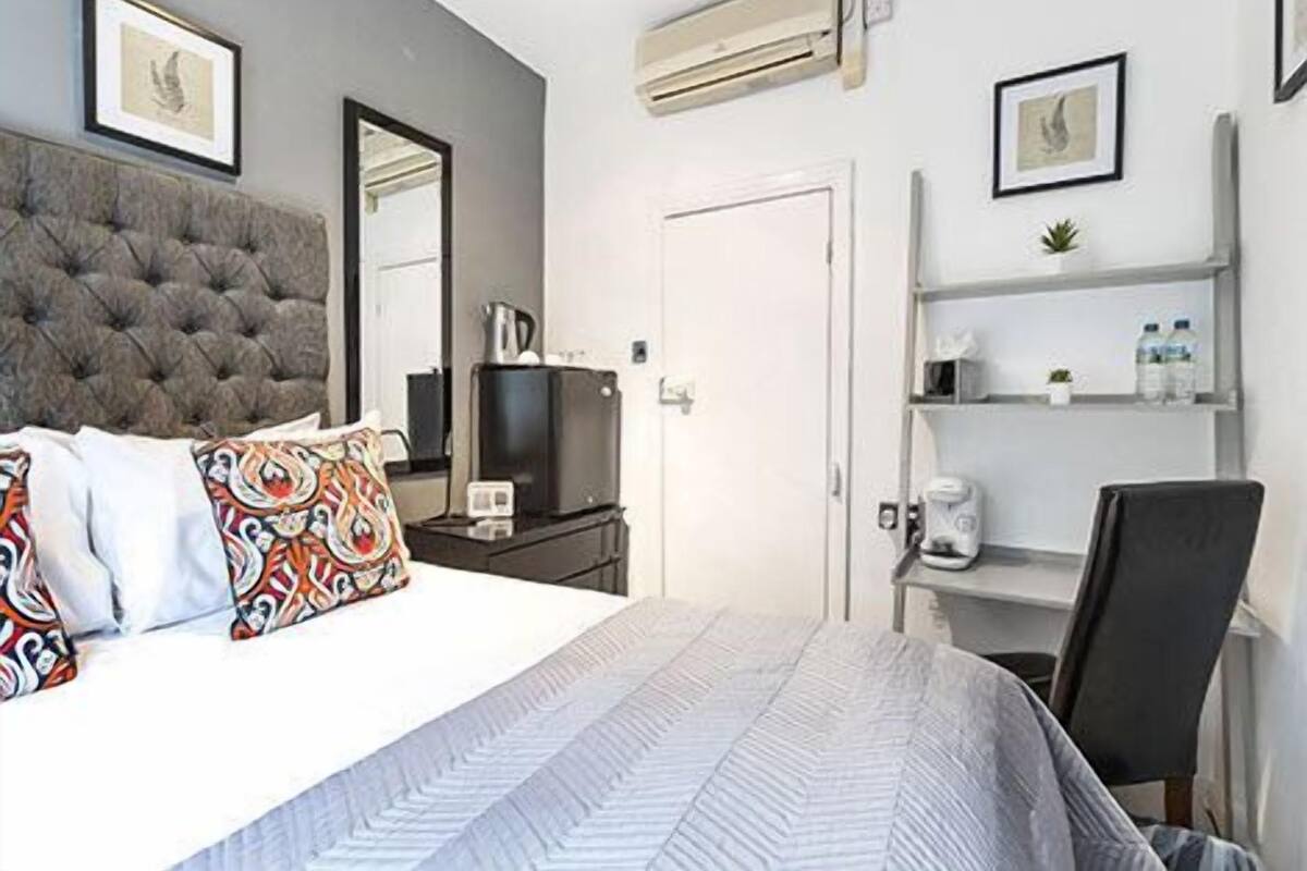 A bedroom with a tufted headboard, a bed with patterned pillows, a television, a coffee maker, a chair, and a shelf with toiletries.