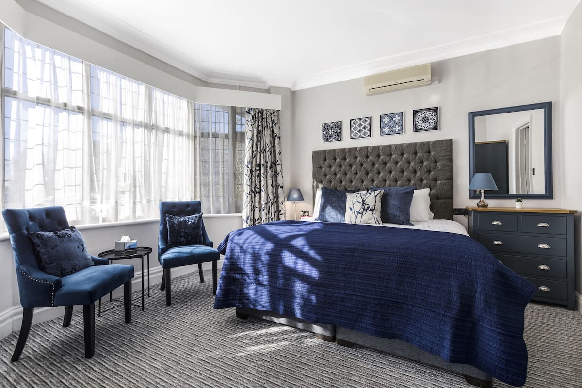 A bedroom with a large bed, two armchairs, a nightstand, and a window with sheer curtains.