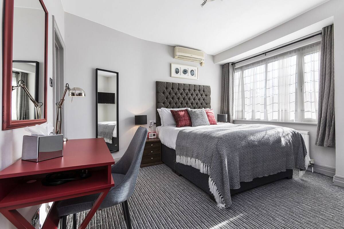 A hotel room with a bed, a desk, a chair, a large mirror, and a window with curtains.