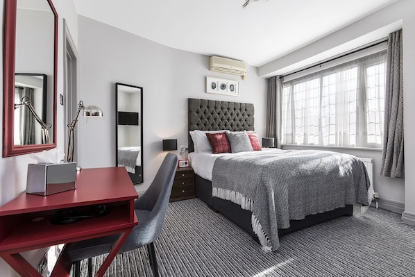 Habitación de hotel con cama, escritorio, silla, un espejo grande y ventana con cortinas.