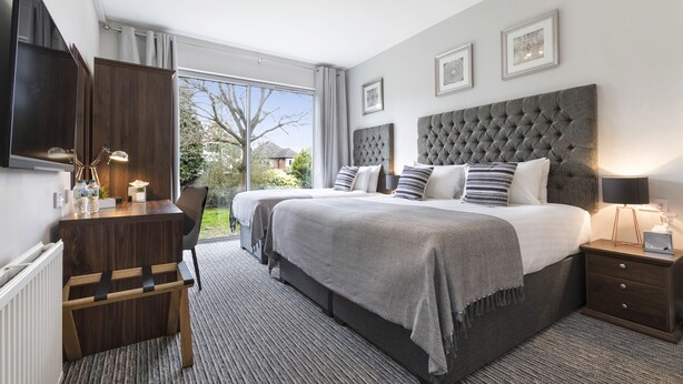 A modern bedroom with a large bed, a desk, and a chair. There is a window with a view of trees and a building outside.