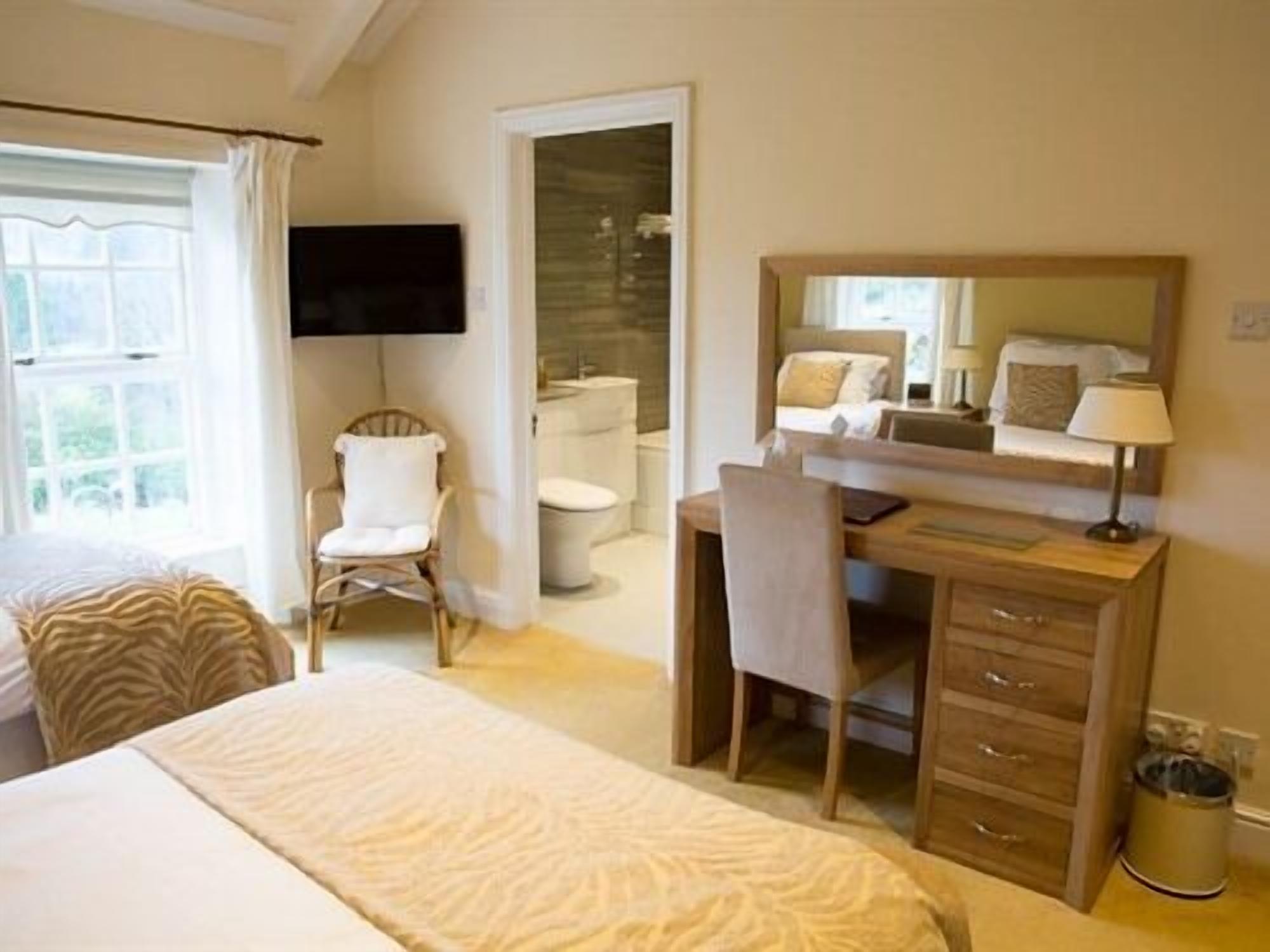Un dormitorio con cama, escritorio, silla, televisor y baño visible a través de una puerta abierta.