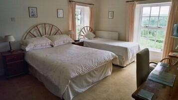 A bedroom with two single beds, a wooden nightstand with a lamp, a wooden desk with a chair, and a window with curtains.