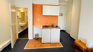 A compact kitchen with a countertop, sink, and a small refrigerator. A trash bin is placed near the sink, and a wooden bench is visible in the corner. - City Apartment Hotel Hamburg (Hamburg)