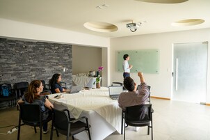 Un salón de conferencias con una mesa blanca, sillas negras, un proyector y una pizarra.