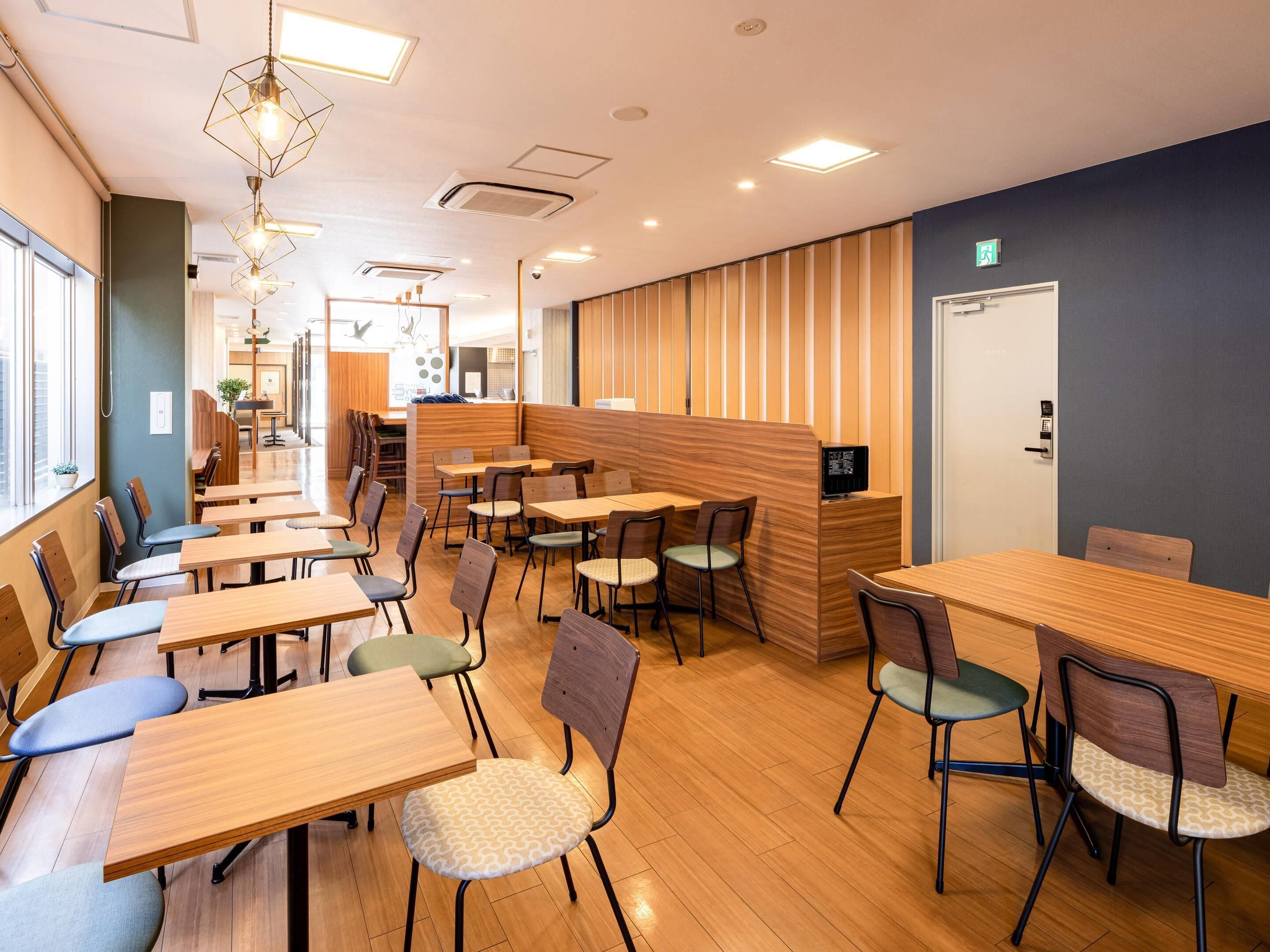 A modern cafe with wooden tables and chairs, a counter with a computer, and a view of the city through large windows.