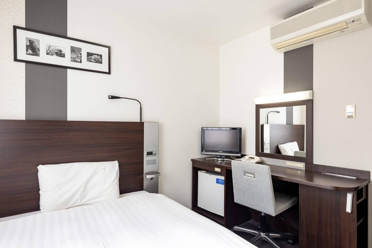 Une chambre d’hôtel avec un lit, un bureau équipé d’un ordinateur, une chaise, une lampe de bureau, un miroir et une photo encadrée accrochée au mur.