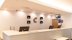 A reception desk with two laptops, a flat-screen TV, and a wall-mounted clock. - Comfort Hotel Shin Yamaguchi (Yamaguchi)