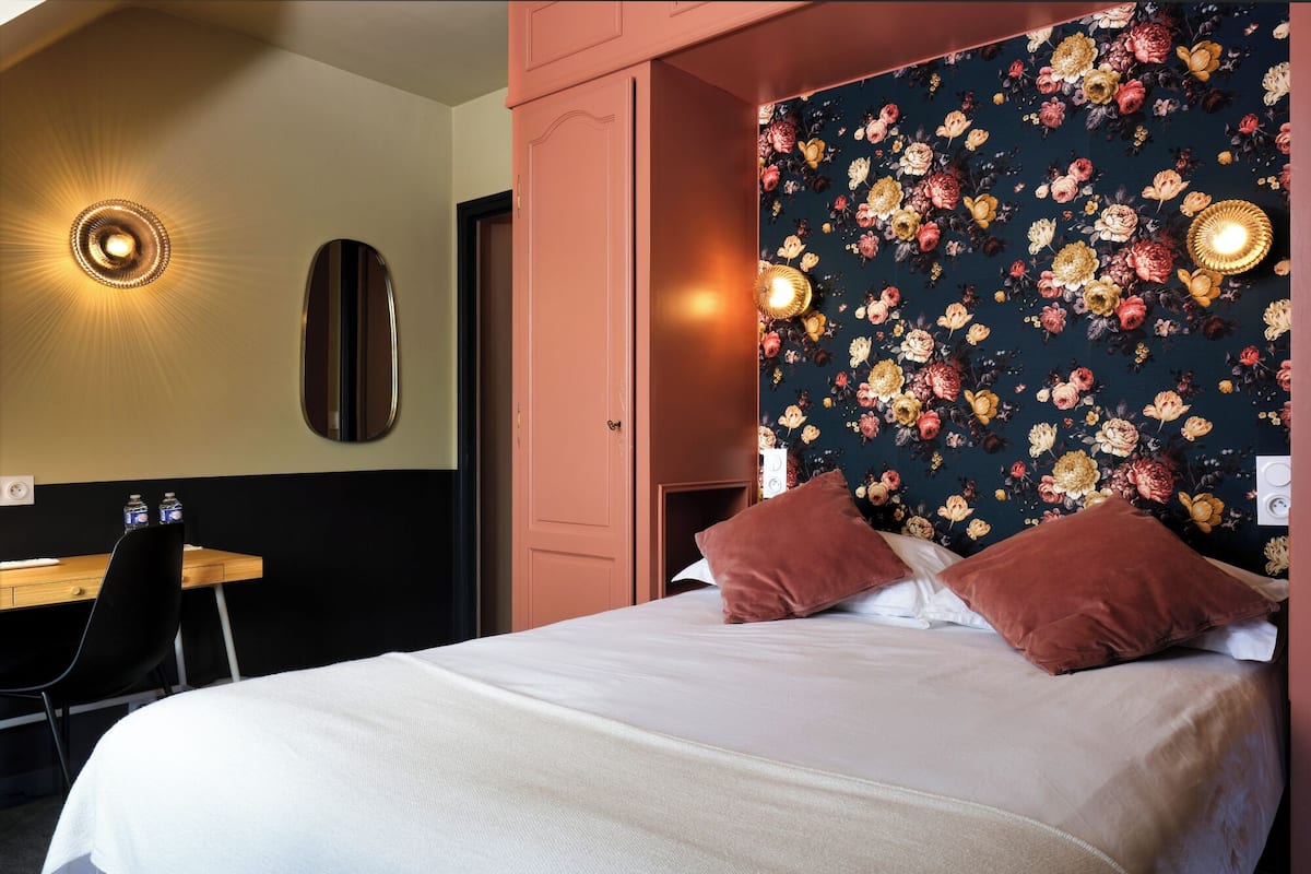 A hotel room with a bed, two pillows, a desk with a chair, a mirror, and a wall with floral wallpaper.