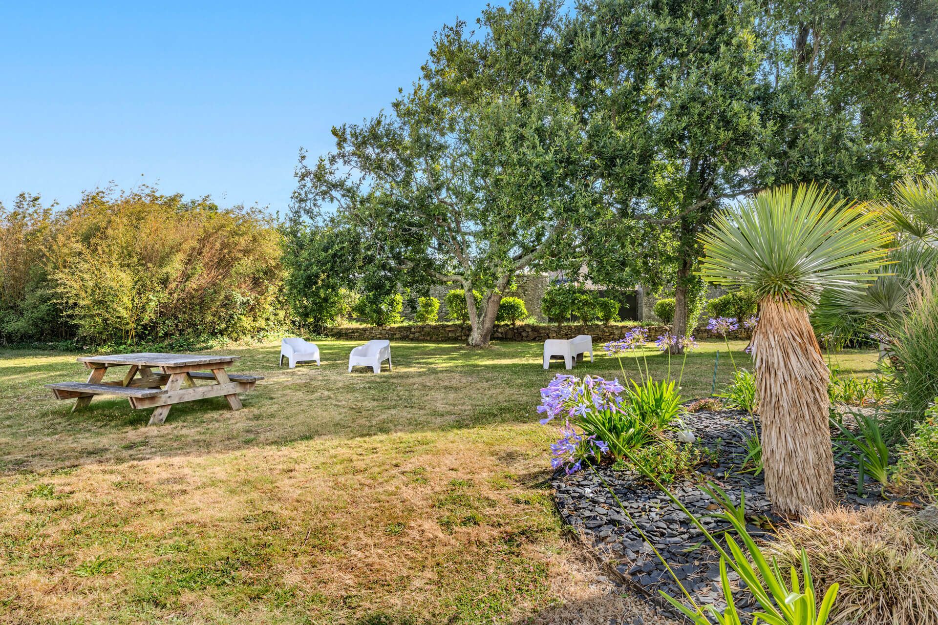 A picnic area with a wooden table and four white plastic chairs, surrounded by a grassy lawn and various plants.