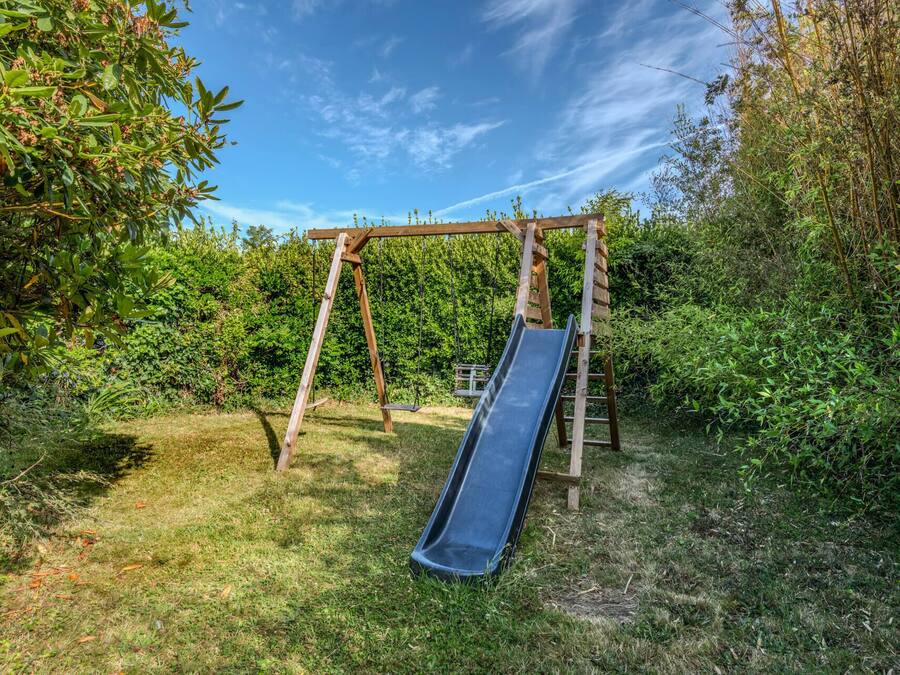A wooden swing set with a slide in a grassy area surrounded by dense foliage.