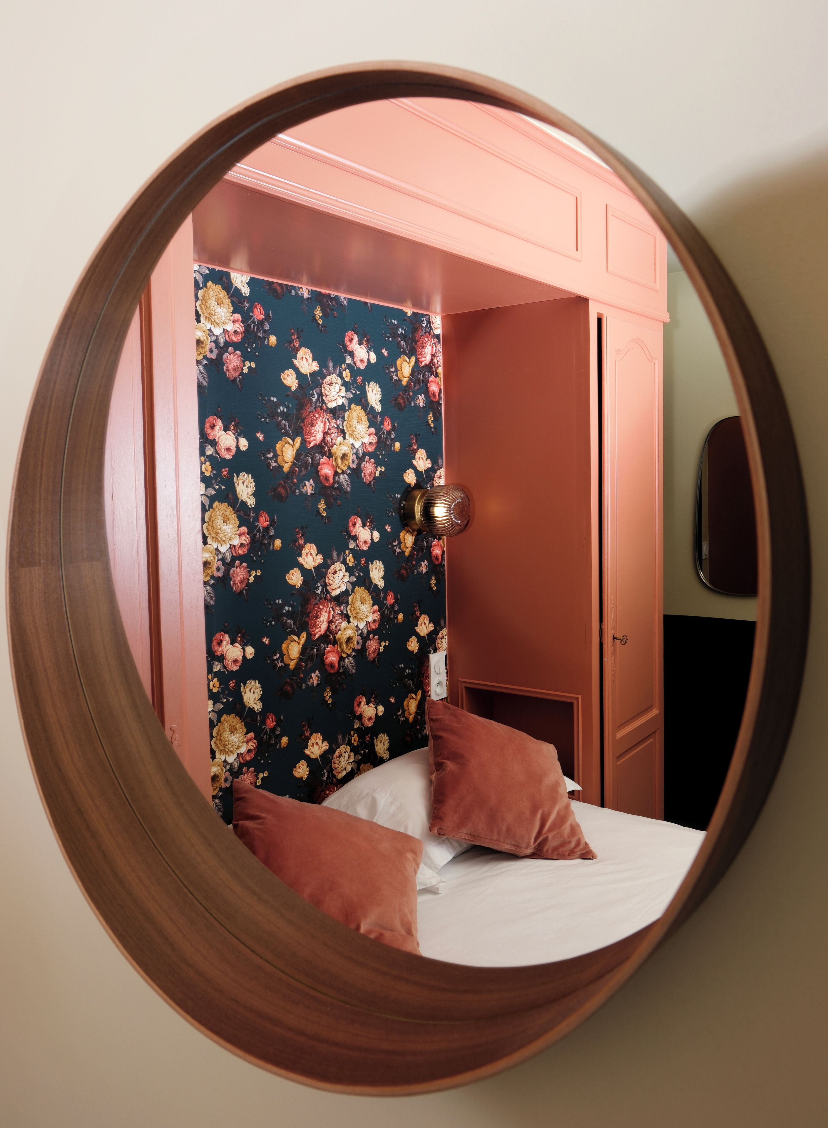 Un espejo circular que refleja un dormitorio con papel tapiz floral, una cama con dos almohadas y una puerta.