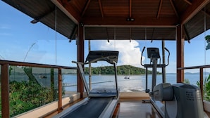 A covered outdoor area with a treadmill and elliptical machine, overlooking a body of water with boats and a forested island. - Chandara Resort & Spa Phuket (Pa Klok)