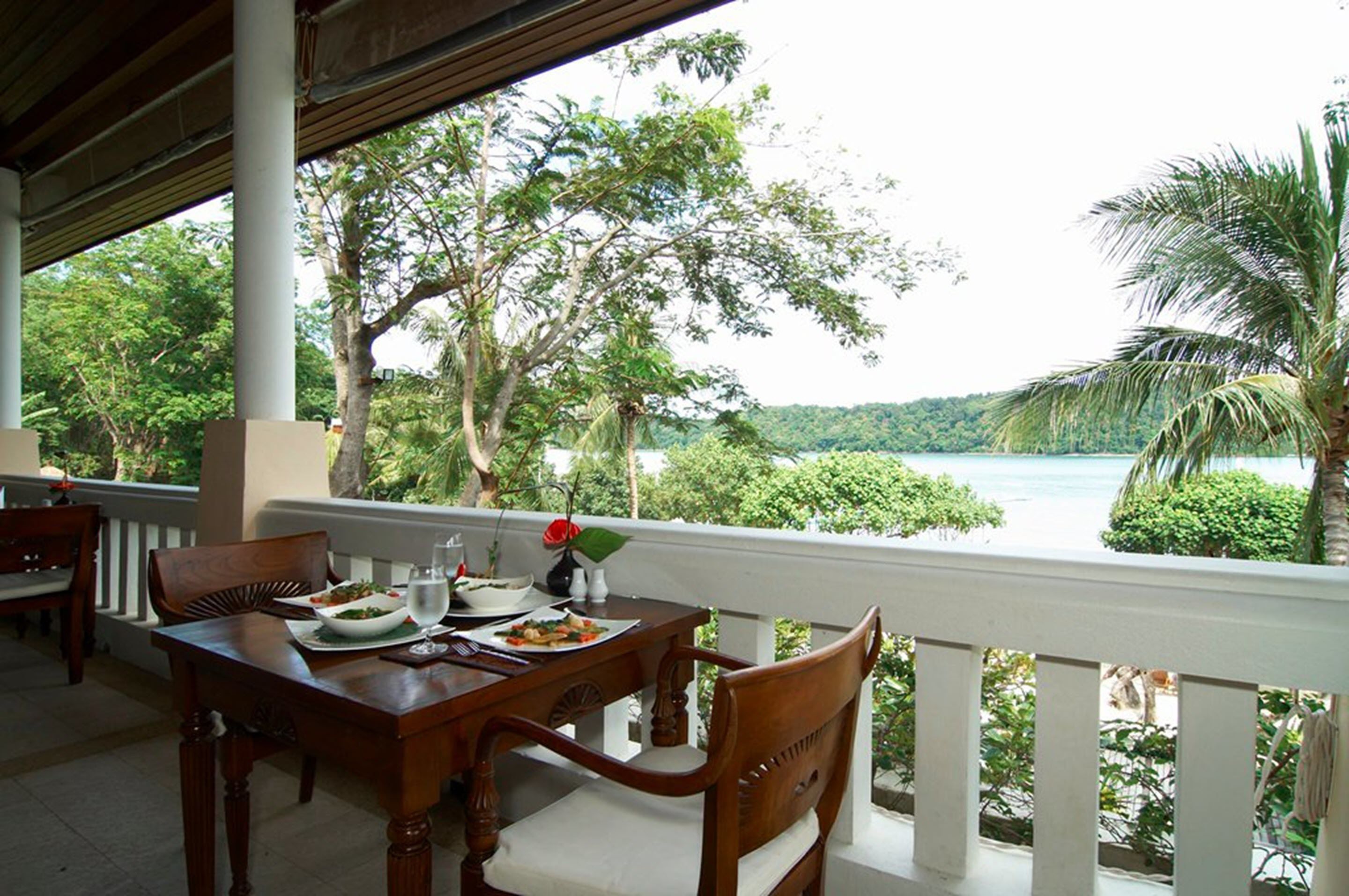A wooden dining table with chairs, a white railing, and a view of a lush green landscape with a body of water and palm trees.