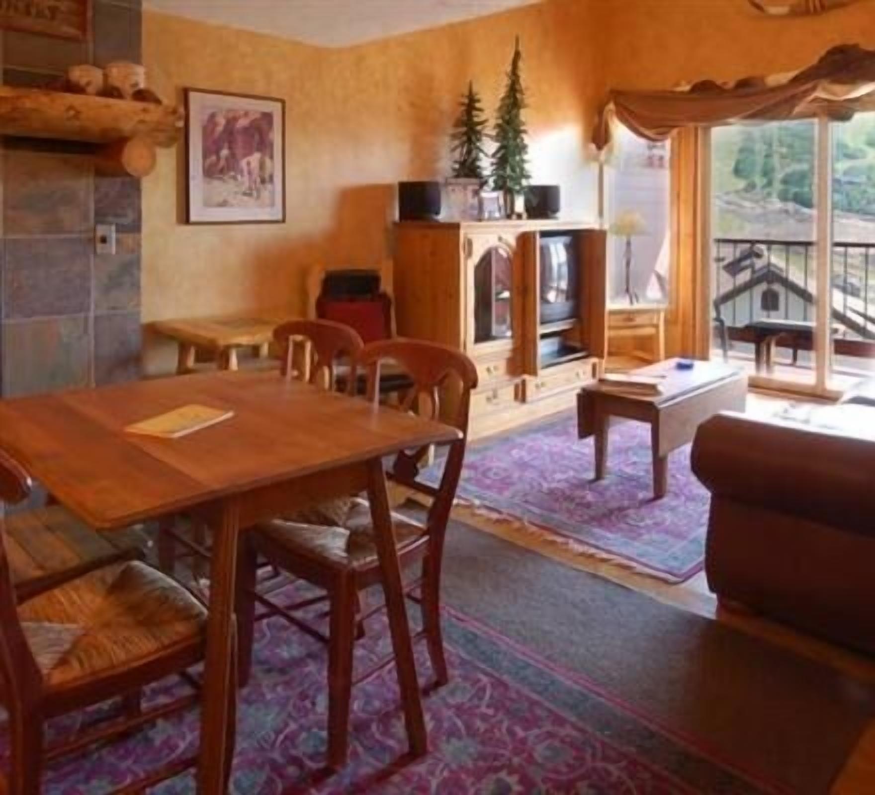 Un salon chaleureux avec une table à manger en bois, des chaises, un canapé et un meuble pour la télévision. Une fenêtre offre une vue sur une maison et un balcon.