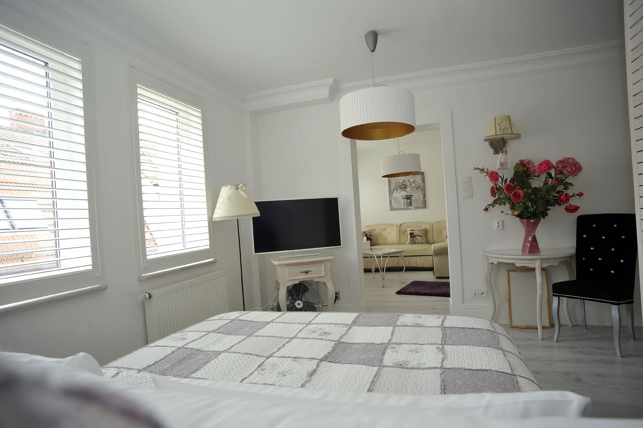 Et soveværelse med seng, fjernsyn, ventilator, lampe, stol, bord med blomster og en sofa i baggrunden.