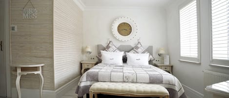 Un dormitorio con cama, mesitas de noche, un banco, un reloj de pared y una ventana con persianas.