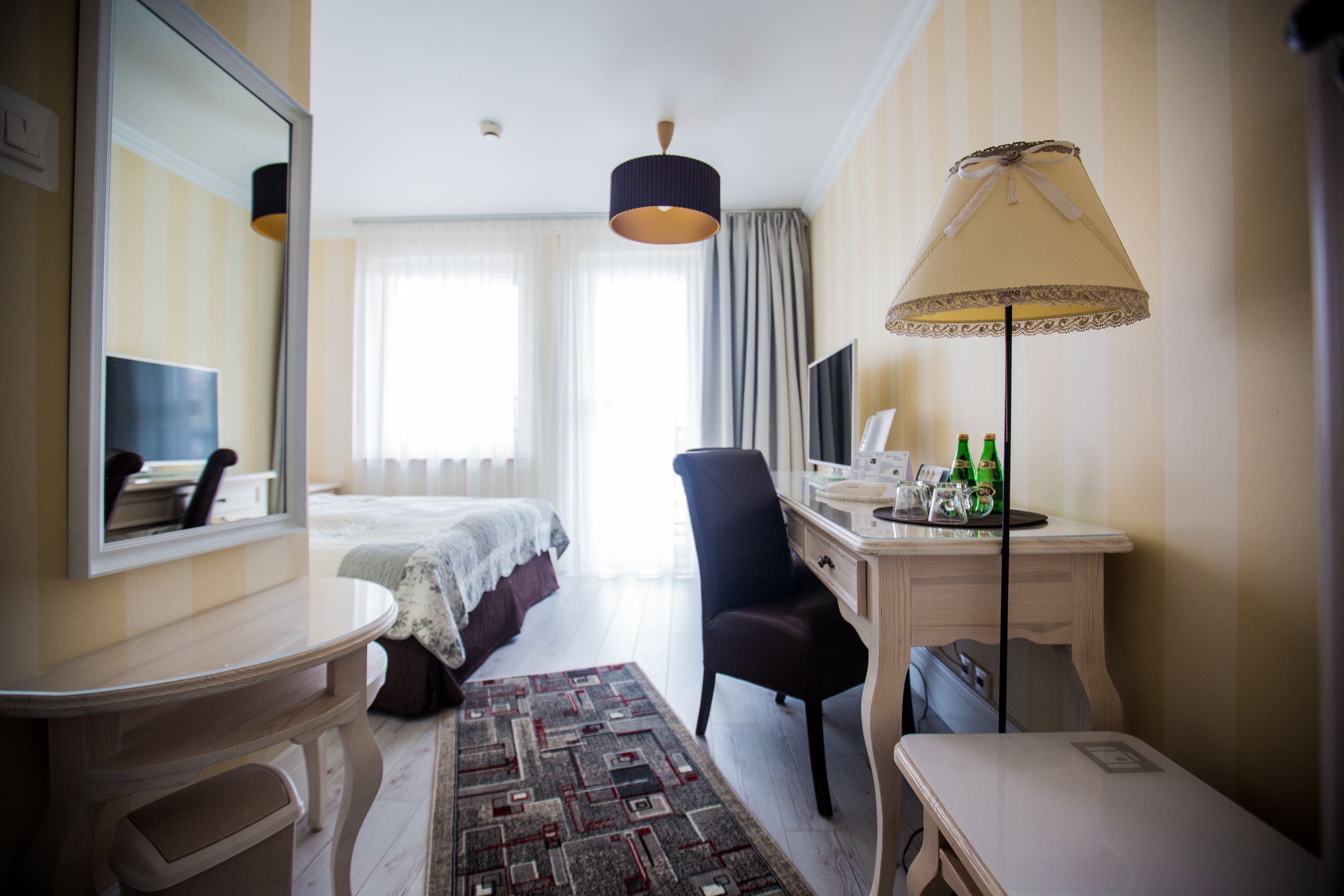 A hotel room with a bed, a desk, a chair, a desk lamp, a mirror, and a television.
