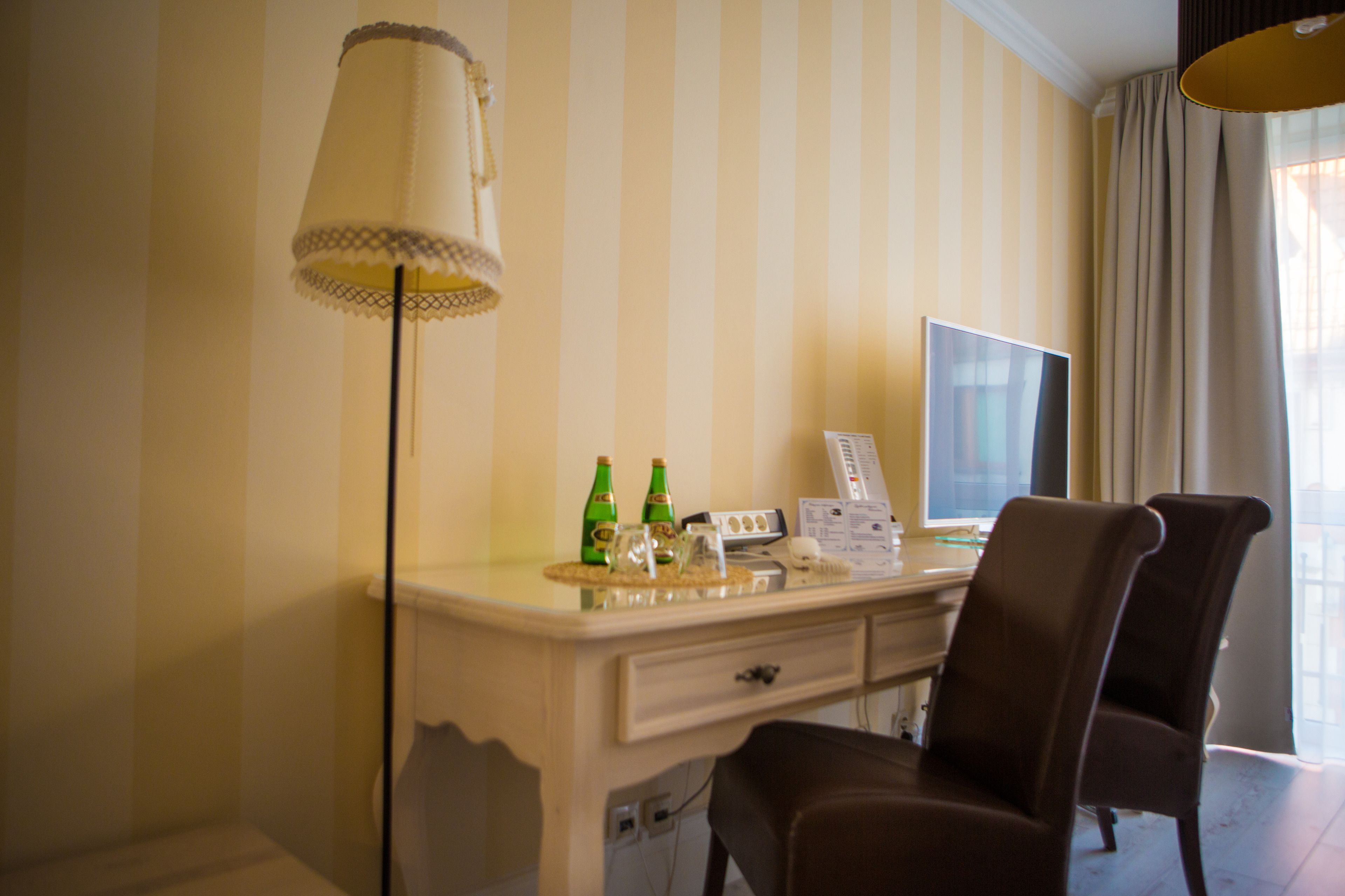 A hotel room with a striped wallpaper, a desk with a computer, two chairs, a lamp, and a window with curtains.
