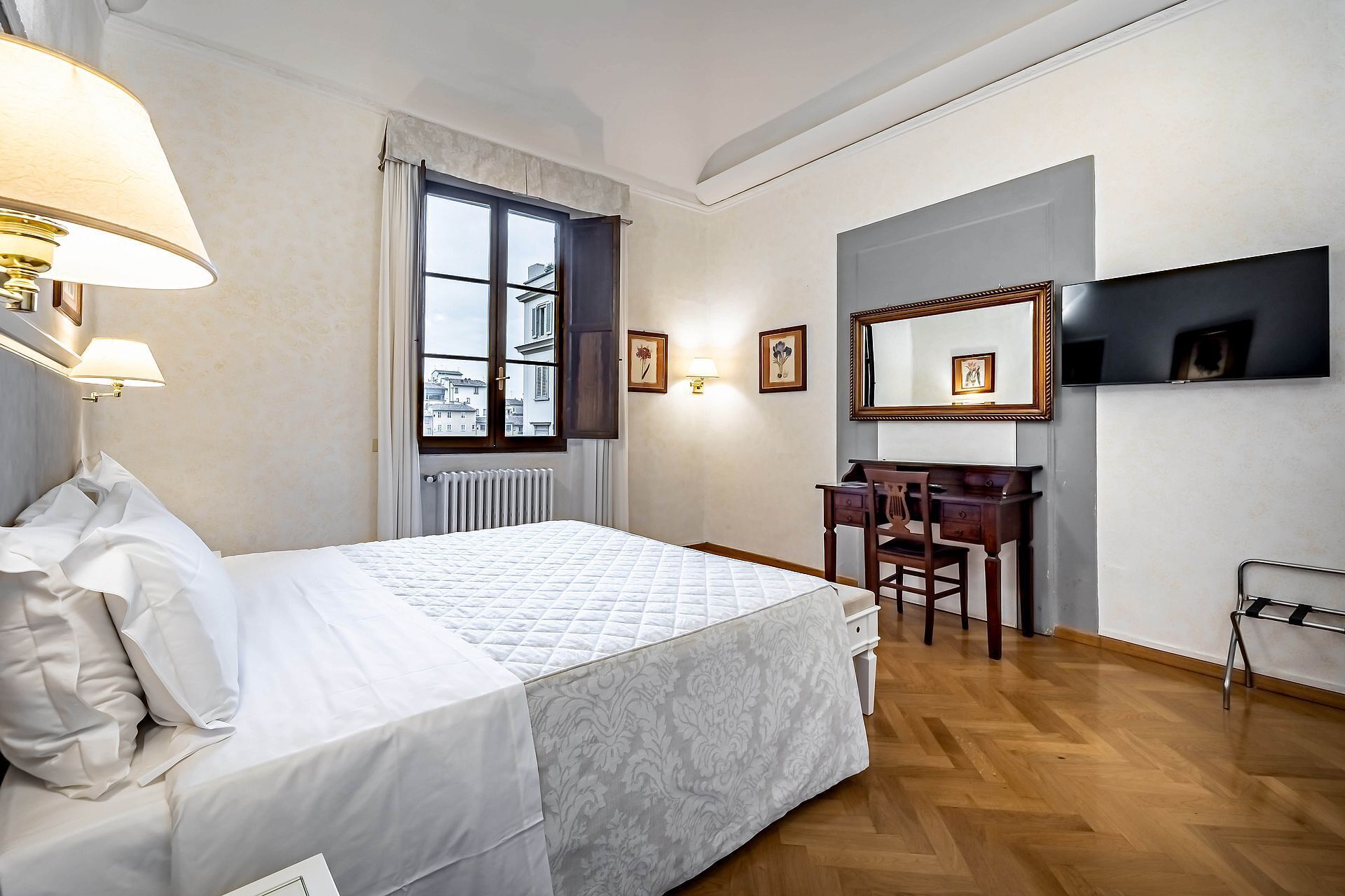 Habitación de hotel con cama, escritorio, silla, radiador, ventana con cortinas y marcos de cuadros en la pared.