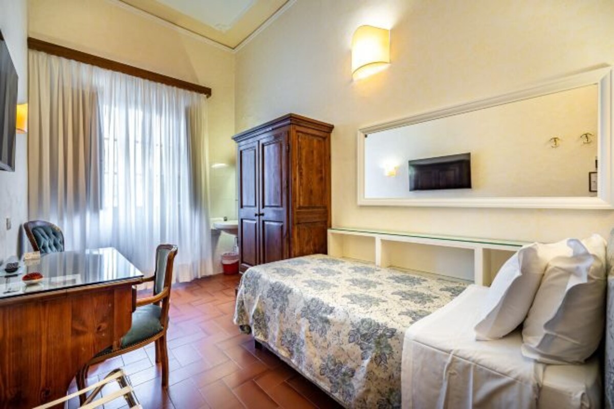 A bedroom with a bed, a desk, a chair, a wardrobe, a TV, and a window with curtains.