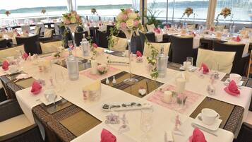 A well-set dining area with tables arranged for guests, featuring elegant table settings, floral centerpieces, and a scenic view of the water.