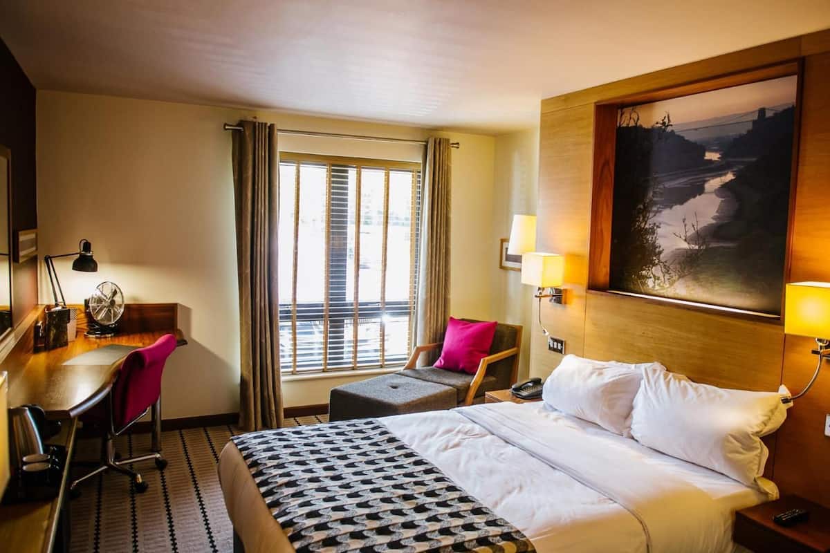 A hotel room with a bed, desk, chair, and a large window with curtains.