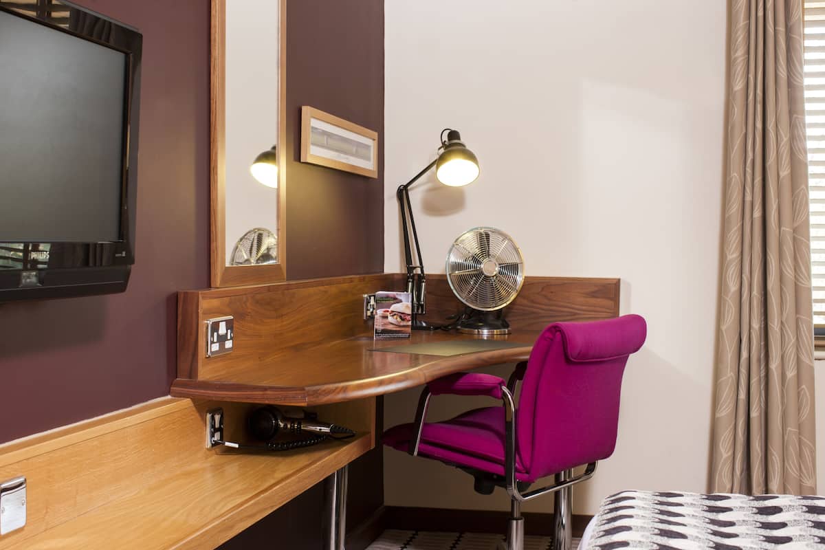 A hotel room with a wooden desk, a pink chair, a television, a desk lamp, a fan, and a bed with patterned bedding.