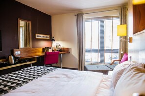 A hotel room with a bed, a desk with a chair, a window with curtains, and a view of the outside. - Lodge at Bristol (Bristol)