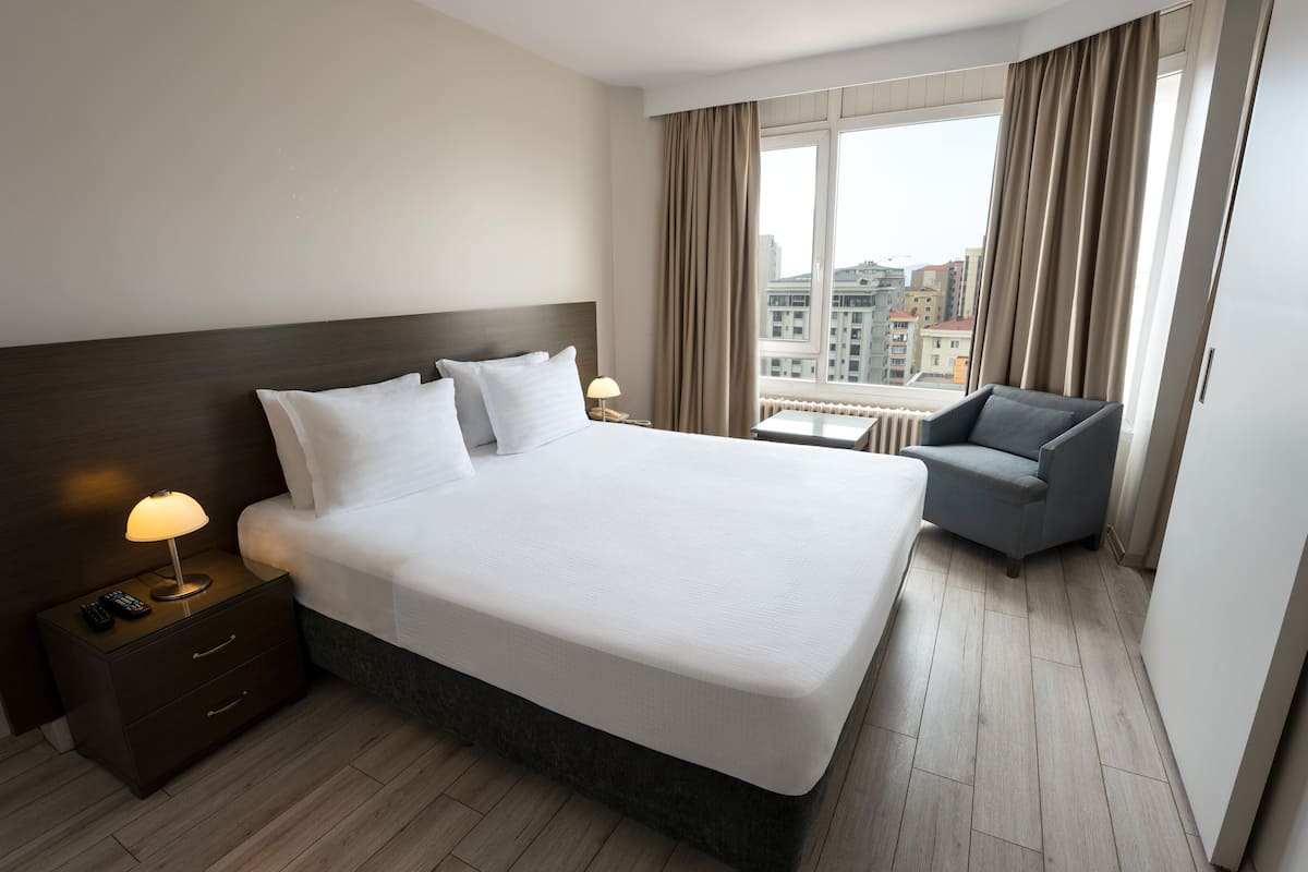 A hotel room with a large bed, a nightstand with a lamp, a chair, and a view of the city through the window.
