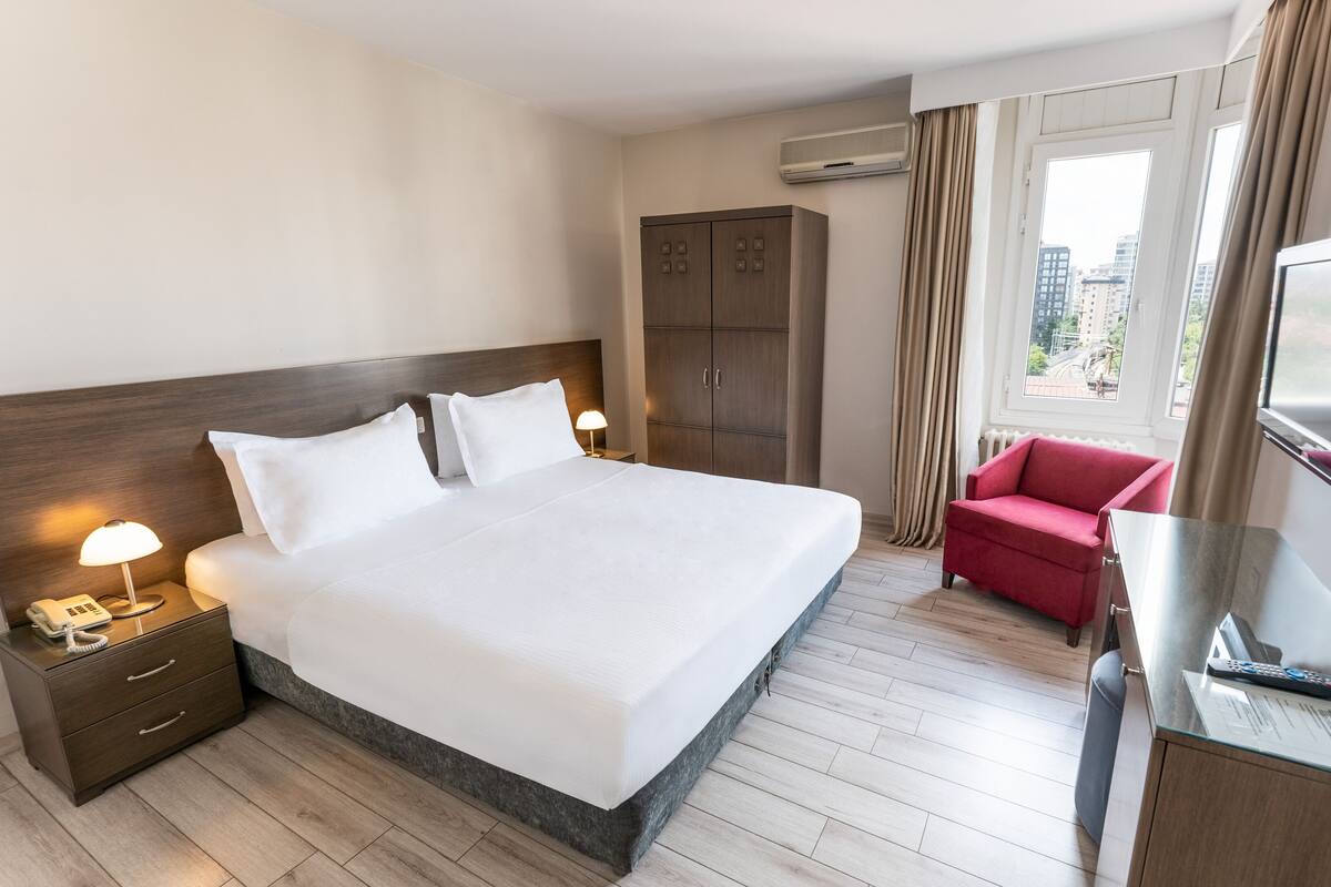 A hotel room with a large bed, a wardrobe, a desk with a TV, and a red chair.