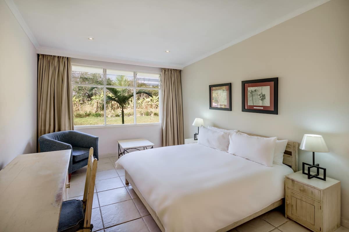 A hotel room with a large bed, a chair, a small table, and a window with curtains. There are two framed pictures on the wall.