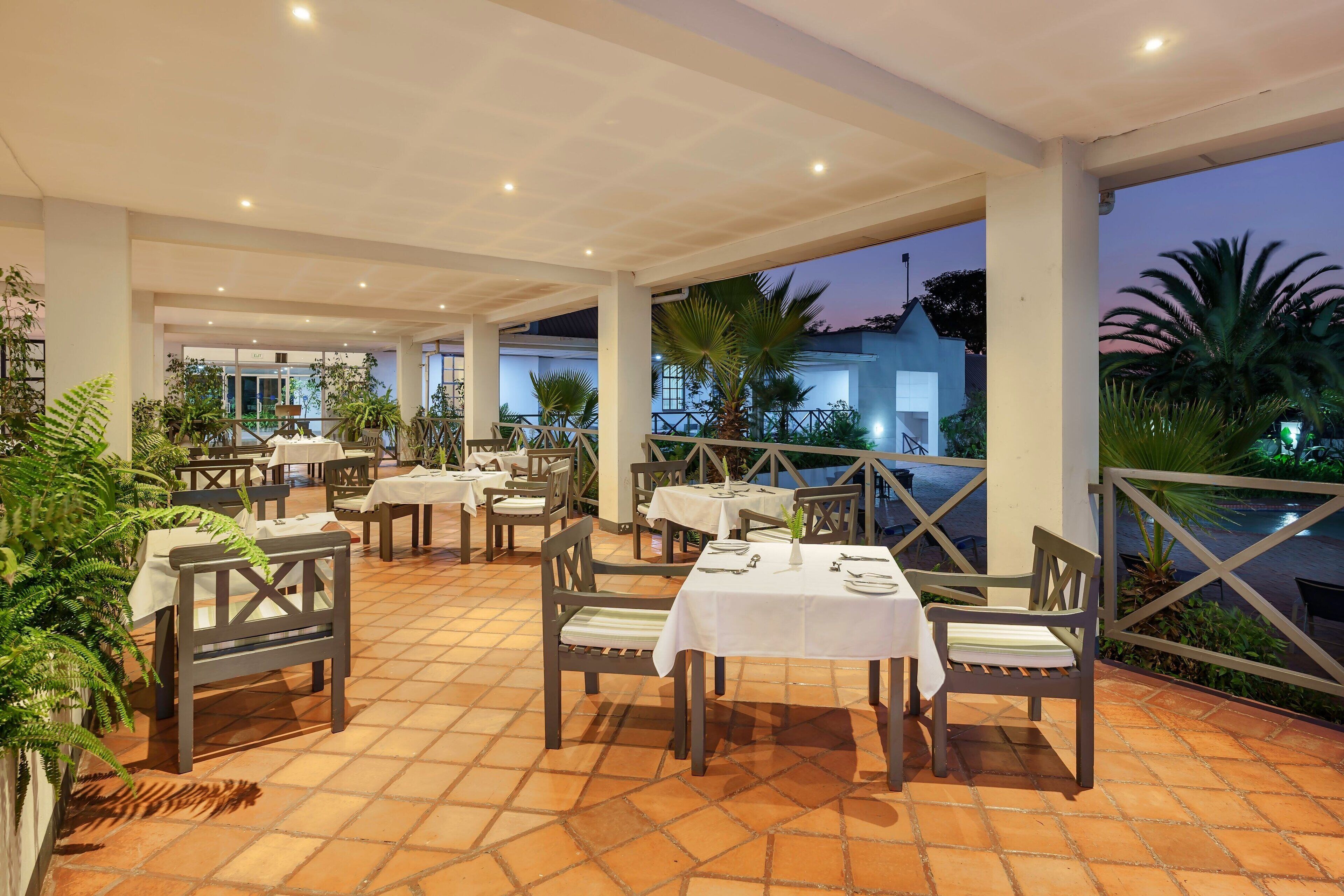 An outdoor restaurant with tables and chairs, surrounded by plants and a view of a building.