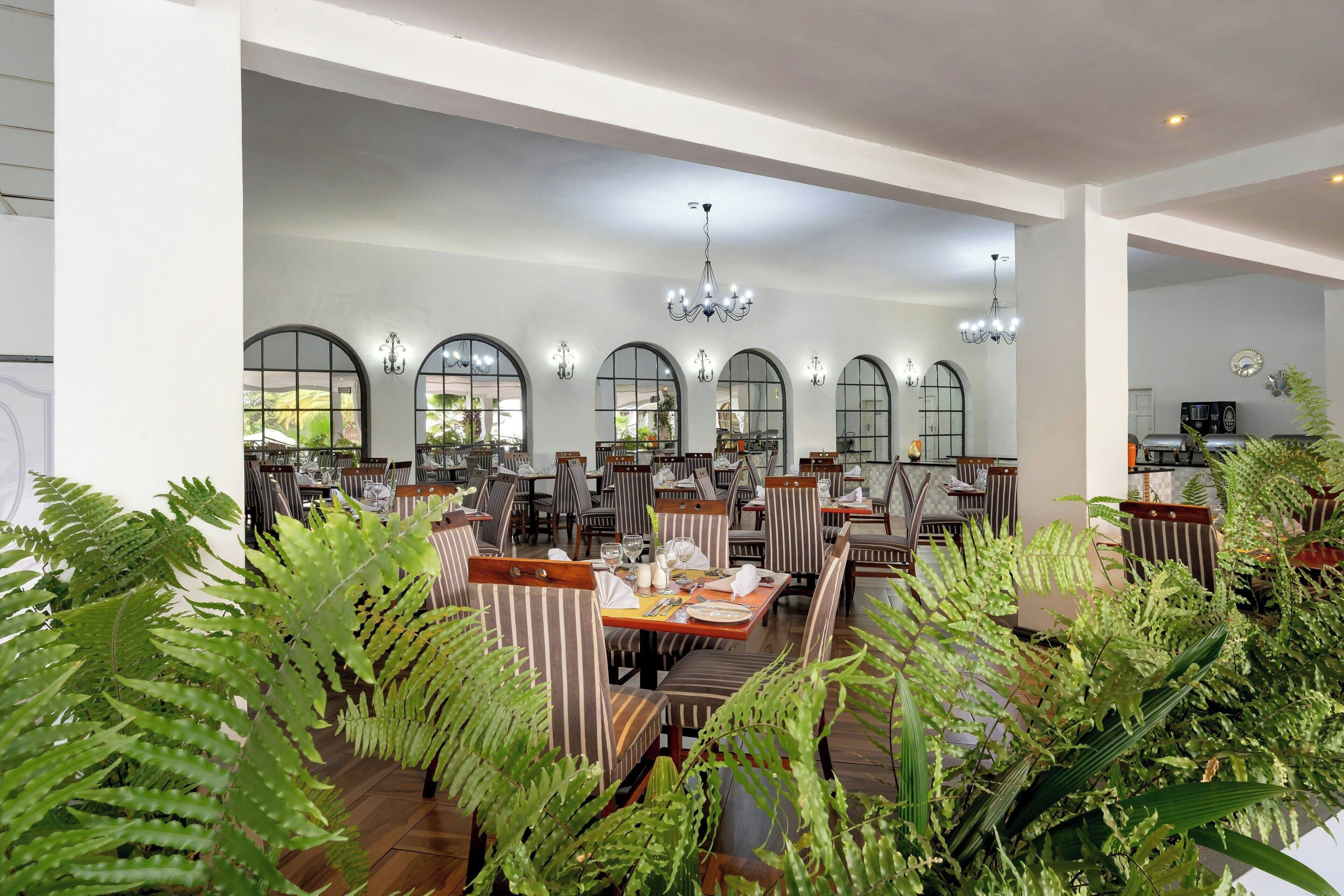Un restaurante espacioso con techos altos, ventanas en arco y una araña. Las mesas están cubiertas con manteles blancos y rodeadas de sillas a rayas.