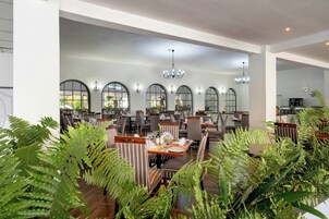 Un restaurante espacioso con techos altos, ventanas en arco y una araña. Las mesas están cubiertas con manteles blancos y rodeadas de sillas a rayas.