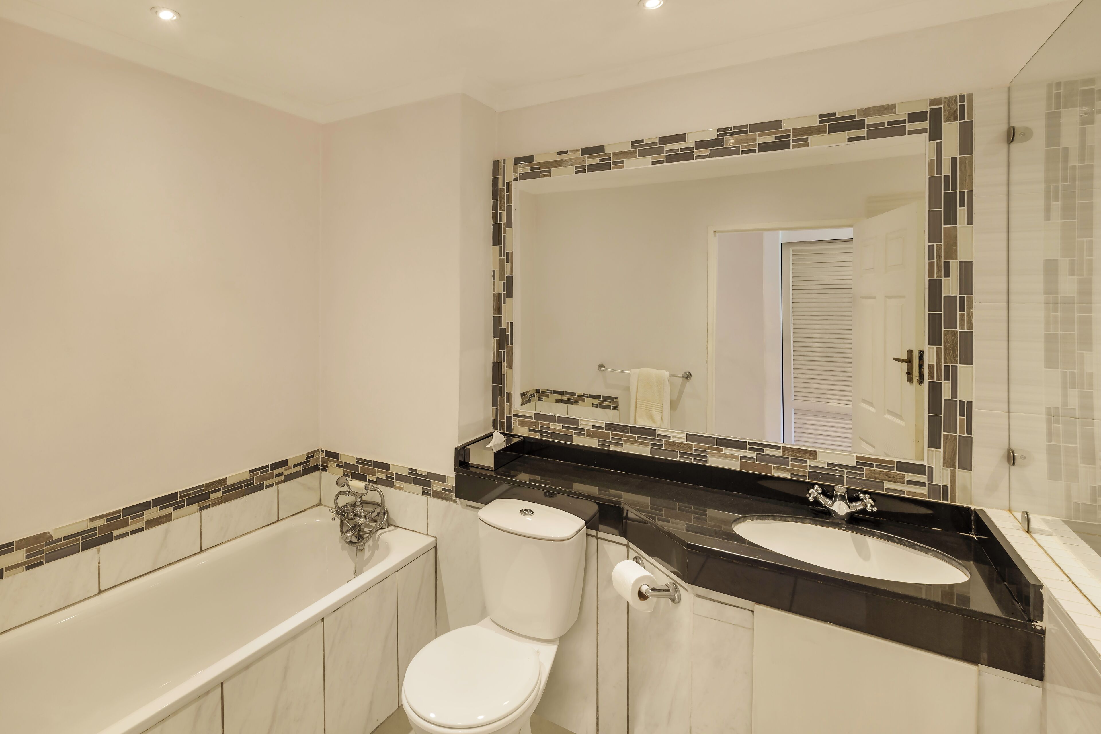 Un baño moderno con bañera, inodoro y lavamanos, que presenta un borde de mosaicos y un espejo grande.